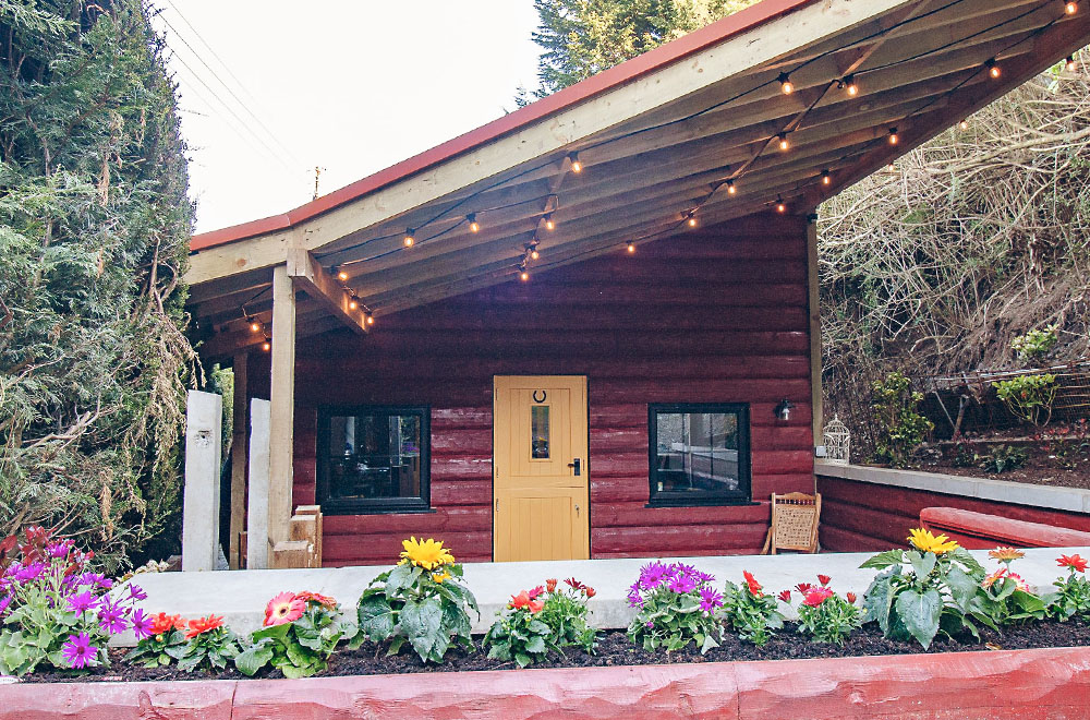 A red stained wood cottage with a yellow door, a slanted roof with string lights and flowers planted in a raised bed in front