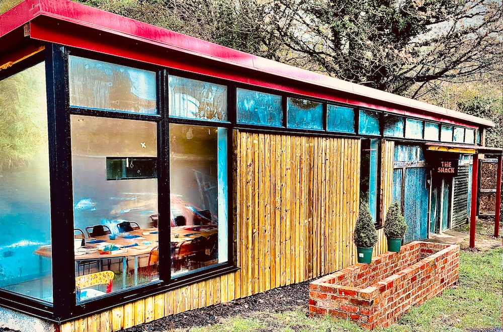 A building with red roof and wood front, there is a sign saying 'The Shack' and large windows showing table and chairs inside. Trees are visible in the background.