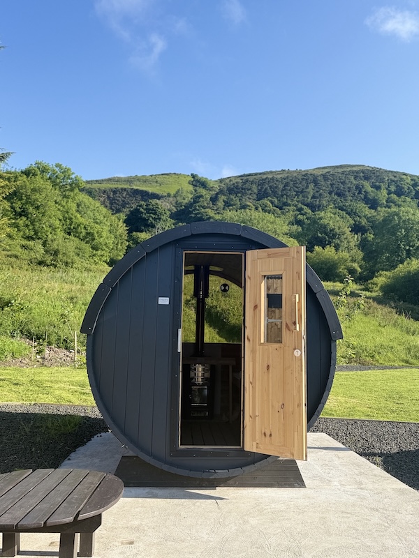 The sauna at Redbarn Cavehill, with blue sky, hills and nature in the background.